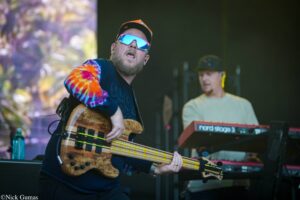 Male bassist playing a wooden bass on stage, wearing a tie-dye sleeve, cap and blue mirrored sunglasses during a live performance.