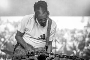 Male guitarist wearing sunglasses and a white t-shirt, playing an electric guitar outdoors at a performance.