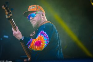 Male guitarist performing on stage, wearing a bright tie-dye shirt and orange camouflage cap with mirrored sunglasses.