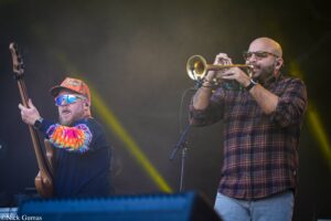 Live concert scene with a trumpeter in a plaid shirt and glasses playing a trumpet beside a bass guitarist in a colorful tie-dye sleeve shirt and hat (stage lighting in the background).