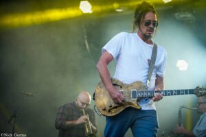 Guitarist wearing sunglasses and a white t-shirt plays a wooden electric guitar on a smoky concert stage with yellow lights in the background.