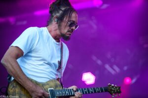 Male guitarist in a light blue shirt playing a wooden electric guitar on a purple-lit stage during a live performance.