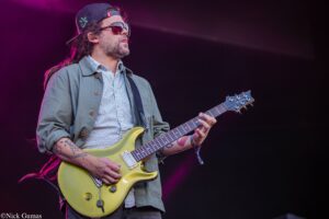 Guitarist performing on stage in a cap and sunglasses, playing a yellow electric guitar.
