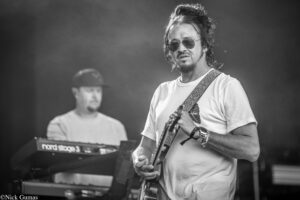 Guitarist wearing sunglasses and a white t‑shirt plays on stage with a patterned strap; a keyboardist is blurred in the background.