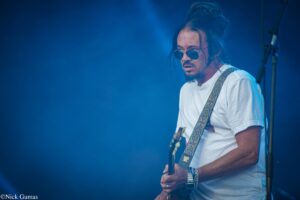 Guitarist wearing sunglasses and a white T-shirt performs on stage under blue lighting, holding a guitar with a patterned strap.