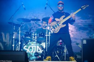 Bass guitarist wearing sunglasses and a colorful cap plays a natural wood five-string bass on a blue-lit stage, with drums and microphones in the background.