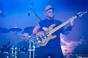Bassist wearing a colorful tie-dye sleeve shows off a wooden bass on stage under blue lighting, with a drummer in the background.