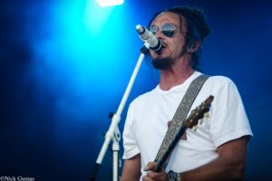 Male guitarist wearing sunglasses sings into a microphone on stage, white shirt and patterned guitar strap visible against blue lighting.