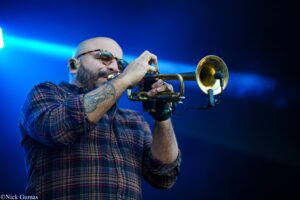 Bald musician in a plaid shirt playing a trumpet on stage under blue lights.