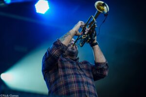 Musician in a red plaid shirt playing a brass trumpet on a blue-lit stage, forearm tattoos visible as he lifts the instrument upward.