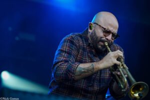 Musician with a shaved head and glasses playing a brass instrument on stage in blue lighting, wearing a plaid shirt.