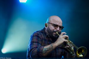 Bald saxophonist wearing plaid shirt playing a saxophone under blue stage lighting.