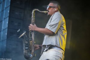 Musician in sunglasses and a light gray shirt plays a saxophone on a dimly lit stage.
