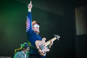 Bassist on stage raises his right index finger while playing a light wood bass guitar.
