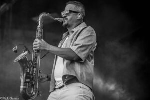 Black-and-white photo of a male saxophonist wearing sunglasses and a jacket, playing on stage outdoors.