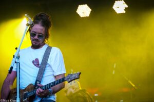 Male guitarist on stage wearing sunglasses and a white T-shirt, playing a wooden electric guitar with a patterned strap against a yellowBackdrop stage lighting.