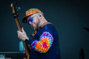 Musician playing a bass guitar on stage, wearing a bright tie-dye shirt, orange camouflage cap, and colorful sunglasses against a dark backdrop.