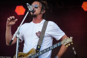 Male guitarist in a white T-shirt with a patterned strap, singing into a microphone on stage with reflective sunglasses on his face.