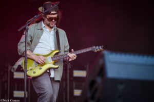Male guitarist wearing sunglasses and a backwards cap, playing a yellow-green electric guitar on stage with a dark backdrop.