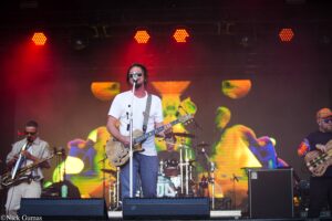 Male guitarist in a white t-shirt and sunglasses sings into a microphone on stage, playing an electric guitar; colorful backdrop and red stage lights behind him.