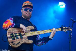 Bass guitarist performing on stage, wearing colorful tie-dye sleeves, a patterned cap, and reflective sunglasses, playing a wooden five-string bass under blue stage lights.