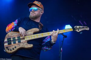 Bass guitarist performing on stage with tie-dye sleeves and an orange cap, playing a striped wooden bass under blue stage lights.
