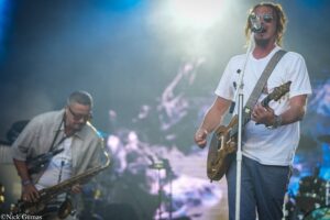 Guitarist in a white T-shirt and aviator sunglasses sings into a microphone while playing an electric guitar; a saxophonist plays in the smoky, blue-lit stage background.