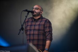 Male singer with beard, bald head, and glasses, performing into a microphone on stage in a red plaid shirt.