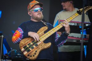 Bearded bassist wearing a camo hat and pink sunglasses, playing a natural wood bass on a live stage.