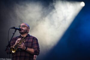 Male musician singing into a microphone on stage while holding a brass trumpet, wearing glasses and a red plaid shirt amid stage smoke and blue lighting