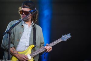 Guitarist performing on stage, wearing a cap and orange sunglasses, playing a yellow electric guitar by a mic stand.