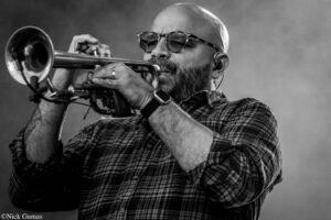 Bearded man wearing glasses and a plaid shirt plays a trumpet, close-up against a dark background (black and white).