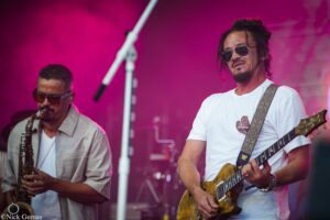 Two musicians perform on a stage bathed in pink light; a saxophonist on the left and a guitarist wearing sunglasses on the right.