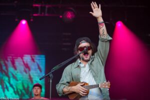Musician on stage with guitar raises right hand amid pink spotlight and dark backdrop, wearing sunglasses and a cap.