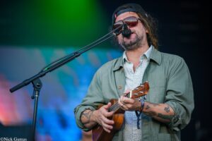 Male musician wearing sunglasses and a backwards cap sings into a microphone while playing a ukulele on stage at a concert.