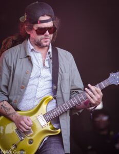 Guitarist wearing a backward cap and sunglasses, playing a yellow electric guitar on stage.