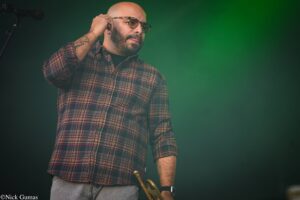 Male musician on stage wearing a plaid shirt and sunglasses, adjusting an ear monitor while holding a brass instrument (trumpet/trenchet) at his side against a green backdrop.