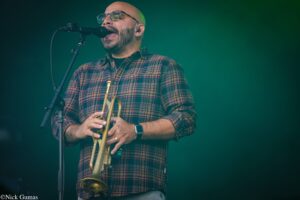 Male musician with a plaid shirt sings into a microphone while playing a brass trumpet on stage.