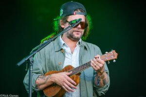 Male musician playing a wooden ukulele on stage, wearing a backward baseball cap, sunglasses, and a green-lit backdrop.
