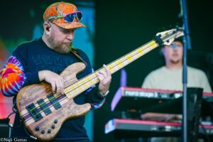 Bass guitarist playing a wood-grain bass on stage, wearing an orange camouflage cap and tie-dye sleeves.
