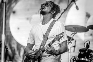 Male guitarist wearing sunglasses and a white t-shirt plays a patterned strap electric guitar on a stage, with drums in the background (black-and-white photo).