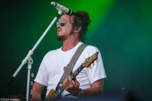 Male guitarist wearing sunglasses and a white T‑shirt, playing a patterned‑strap electric guitar on a green-lit stage.