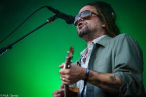 Male guitarist singing into a microphone during a live performance, wearing sunglasses and a patterned shirt against a green backdrop.