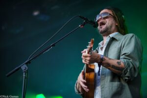 Male musician singing into a microphone while playing an acoustic guitar on stage, wearing sunglasses and a patterned shirt; green stage lighting on him.
