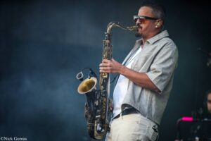 Male saxophonist wearing sunglasses and a light gray jacket, playing a saxophone on a dim stage backdrop.