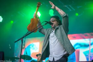 Stage musician on a concert stage, raising a small wooden guitar and singing into a microphone under green lights