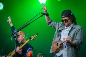 Two musicians perform on a neon-green stage; the foreground guitarist raises a peace sign and plays an acoustic guitar while wearing sunglasses and a cap.