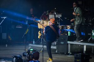 Musician in colorful cap and yellow-and-black sneakers plays a bass on a dimly lit stage; drummer and guitarist visible in background under blue spotlight