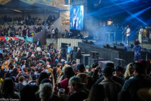 Large outdoor concert crowd facing the stage with musicians performing under blue stage lights and a large screen.