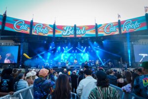 Crowd at an outdoor concert in front of a large colorful stage banner reading Cali Roots, with musicians performing under blue lights.